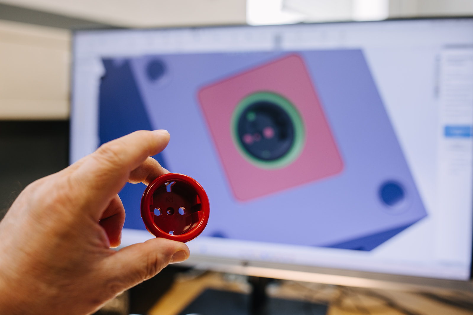 Hand hält rotes Kunststoffteil vor Bildschirm mit CAD-Modell der passenden Form