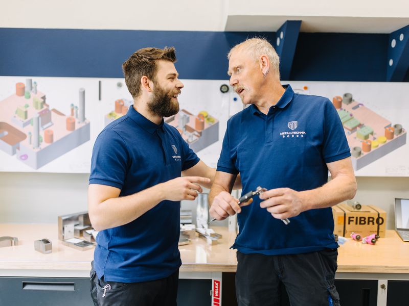 Erfahrungsaustausch im Werkzeugbau: Kollegen prüfen Bauteil mit Messschieber vor CAD-Plänen.